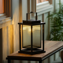 Black solar LED lantern lit on outdoor railing, warm light behind glass panels.