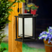 Black solar LED lantern mounted outdoors, warm light shining through front glass.