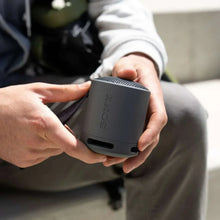 Hands holding a dark gray portable Bluetooth speaker with carry strap and grille top