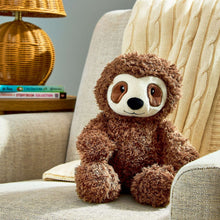 Brown sloth plush toy seated on a chair cushion, white muzzle, curly fur, green tag