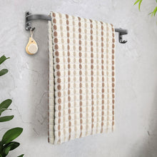Tan bath towel set with dotted grid pattern and fringed ends hanging on a rack