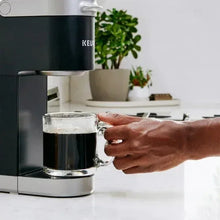 Keurig coffee machine dispensing into glass mug, dark coffee with hand holding mug