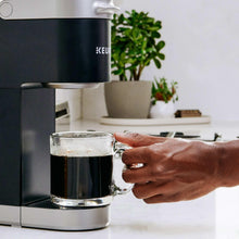 Hand placing a mug under a Keurig machine dispensing dark coffee