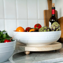 White Staub serving bowl on wooden stand, surrounded by fresh produce and greens