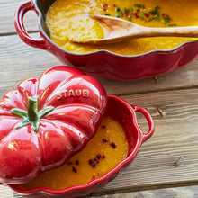 Red tomato cocotte with lid off, showing yellow baked dish inside and embossed STAUB