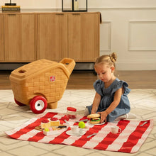 Tan picnic play basket on red wheels, toy food and cups on striped blanket.