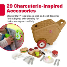 Toy picnic basket with beige body and red wheels next to assorted pretend charcuterie pieces