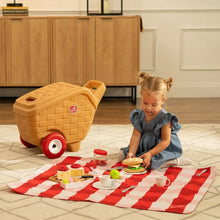 Tan picnic play basket on red wheels beside a toddler arranging toy food on blanket.