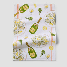 White champagne-themed kitchen towel with bottles, lemons, herbs, and oyster plate print.