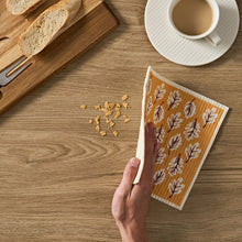 Dishcloth with tan background and brown leaf pattern, hand holding over table