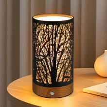 Table lamp with black cylindrical branch-pattern shade and wooden base, warm glow on.