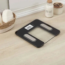 Body composition scale with dark glass top and LCD display on a wooden floor.