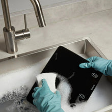 Black digital kitchen scale in sink with water splash, showing touch icons