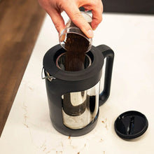 Black cold brew coffee maker with lid open and stainless filter holding coffee grounds