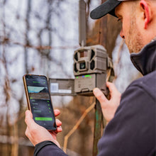 Cellular trail camera outdoors with antenna, shown being configured on a smartphone app