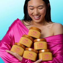 Pack of Tree Hut Tropic Glow firming sugar scrub jars held in arms, golden beige