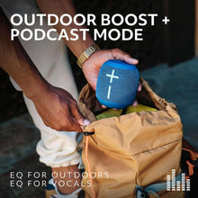 Blue wireless Bluetooth speaker held above an outdoor bag, cross icon on front