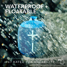 Blue wireless Bluetooth speaker with cross emblem, held with splash of water droplets