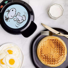 Black waffle maker with Snoopy waffle plus fried eggs and fork on countertop