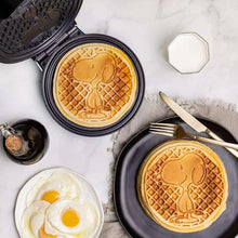 Round Snoopy waffles in open waffle maker with fried eggs nearby