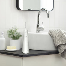 White cordless water flosser handle standing beside a sink faucet on a countertop.