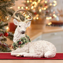 Resting deer figurine in light beige, holding a green berry wreath on its chest.