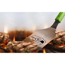 Stainless BBQ spatula with green handle and NFL logo, grilling over open flames