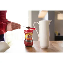 Folgers instant coffee crystals jar near white creamer pitcher on tabletop, hand nearby