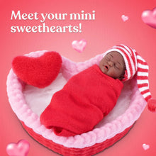 Tiny baby doll in red outfit on pink heart blanket with striped hat