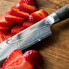 Stainless Damascus chef knife blade with “Bobby Flay” branding on wooden board