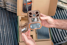 Tan trail camera with open battery compartment being installed by hand