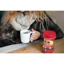 Folgers instant coffee crystals jar on table next to white cup, hand holding lid