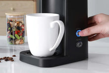 Black matte single-serve coffee maker base with white mug and finger on buttons