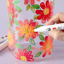 Hand using a paint pen to create colorful flower designs on a clear surface