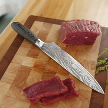 Chef knife with gray Damascus-pattern blade cutting red meat on a wooden board