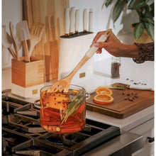 Clear glass cooking pot on stovetop with lid and side handles, herbs and vegetables inside.