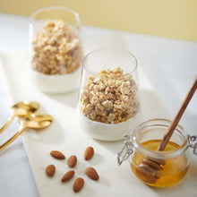 Granola clusters in a glass bowl with honey jar and almonds on white surface