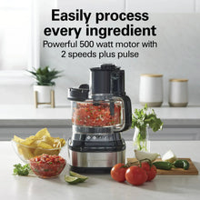 Black food processor with clear bowl and lid on countertop with vegetables and prepared ingredients