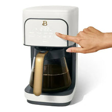 White programmable coffee maker with gold-tone handle and finger pointing at the panel