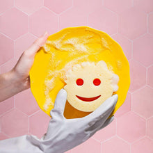 Yellow cleaning sponge with pink scrub face and smiling center, held on pink tiles