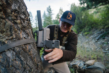 Beige cellular trail camera mounted on a tree while a person adjusts the device.