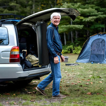 Black folding camping chair in carry bag at outdoor campsite near a vehicle trunk.