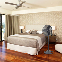 Black pedestal fan with round grille standing on floor in a bedroom setting