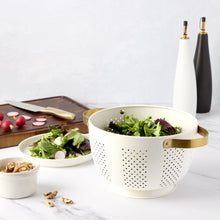 Cream perforated colander with gold handles on countertop beside greens and produce.