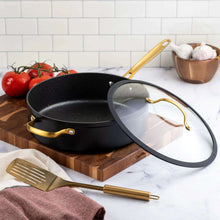 Black non-stick sauté pan with glass lid, gold handles, and utensils