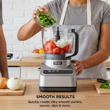 Gray food processor with clear bowl holding chopped carrots and spinach, pouring ingredient