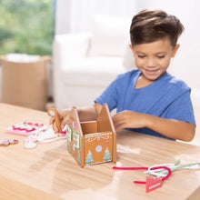 Child lifting a pink wooden gingerbread house with laces and candy-shaped pieces nearby