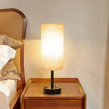 Bedside table lamp with glowing linen shade and black base on nightstand