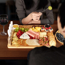 Bamboo cheese board set with white-handled knives and assorted sliced cheeses and fruit