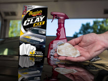 Hand using a white clay bar on car paint with red spray bottle and kit box nearby
